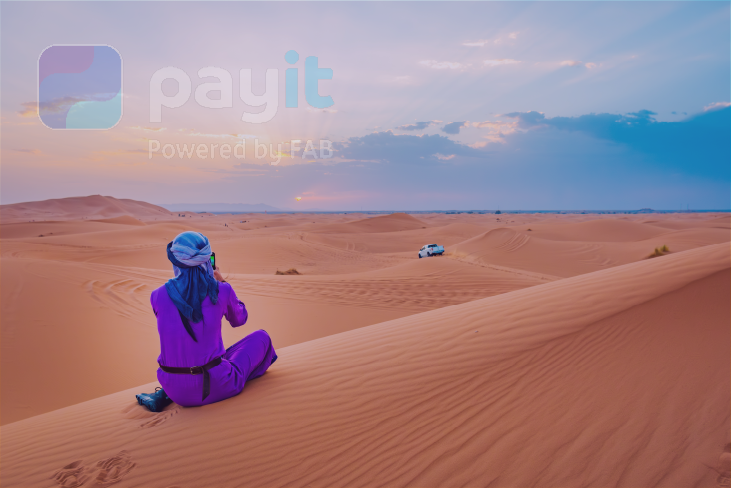 A man in traditional Arab attire sits in the desert, capturing photographs. The image is watermarked with the Payit logo and serves as a banner representing the best camping spots in Dubai, UAE